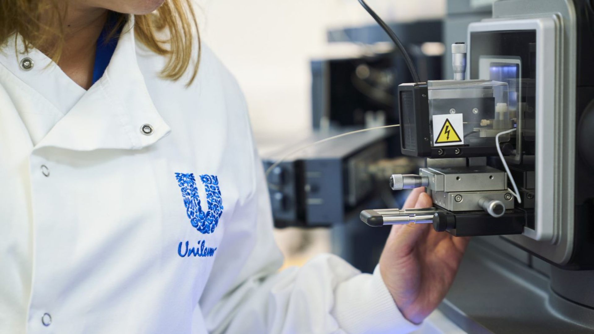 A close-up photo of a scientist in a Unilever-branded white lab coat, looking at a large digital microscope.